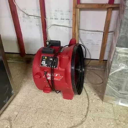 A red air mover running in a room where the lower wall has been opened and plastic sheeting hangs to contain the work area